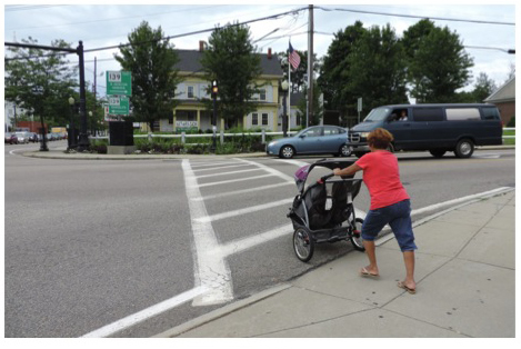 Figure 5, titled “Pedestrian Crossing on Plymouth Street.” This is a photograph of a woman pushing a stroller as she enters the crosswalk on Plymouth Street.